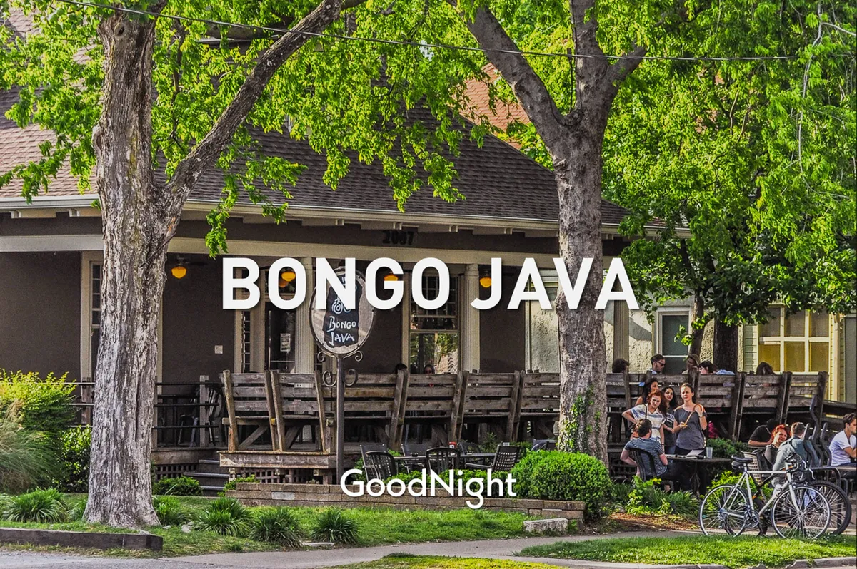 This image captures the vibrant exterior of Bongo Java, featuring a prominent sign, outdoor seating shaded by trees, and people socializing and biking nearby. The casual and friendly atmosphere is evident through the communal seating and relaxed interactions visible in the scene.
