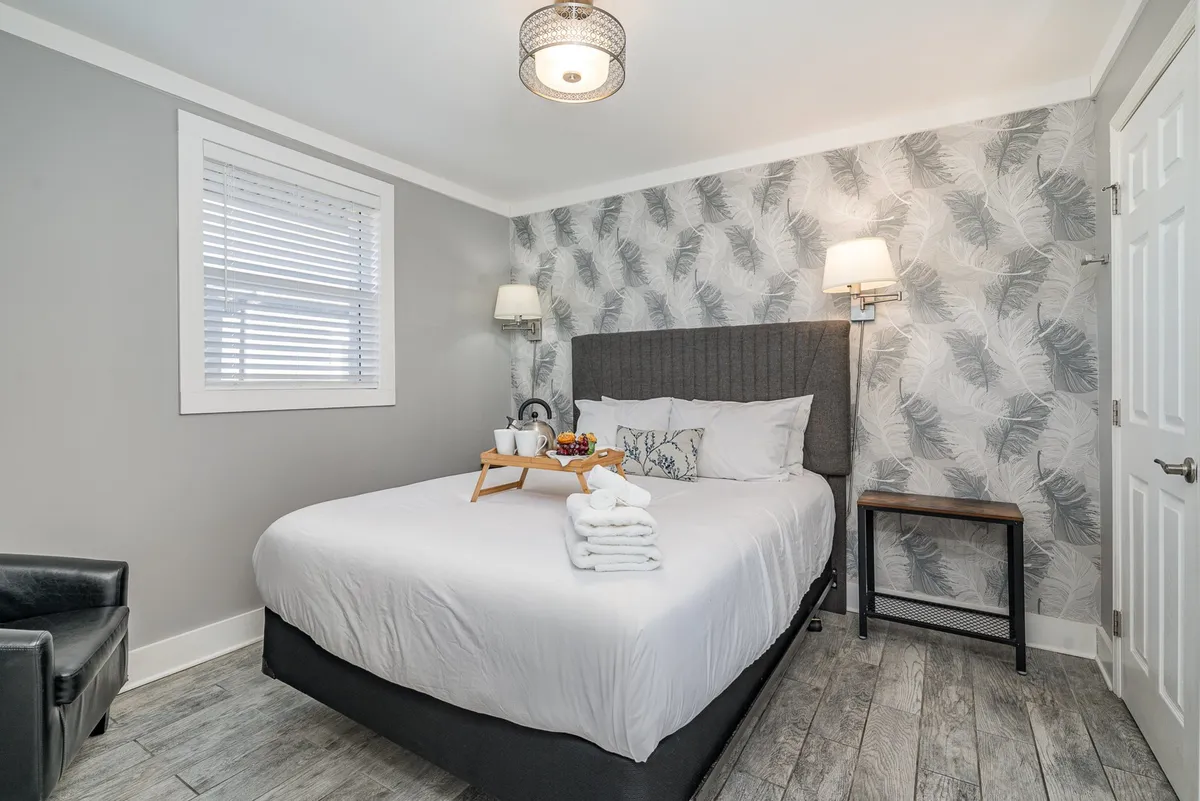 This image features a serene bedroom with a plush bed adorned with decorative pillows. A breakfast tray with fruit and drinks sits on the bed, while leafy wallpaper and an accent chair create a cozy, relaxing atmosphere. The space is softly lit, emphasizing a tranquil, restful ambiance.