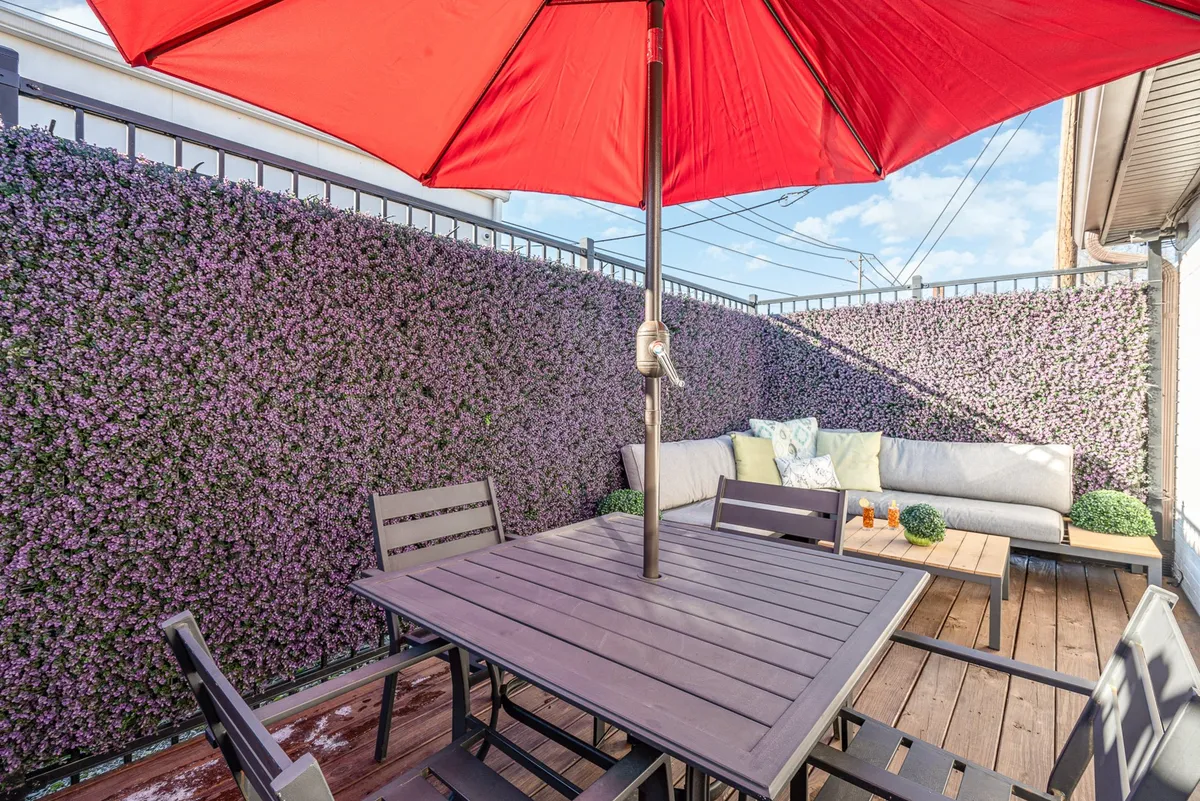 This image features a cheerful outdoor dining area with a red umbrella, comfortable seating, lush greenery on surrounding walls, and a wooden table ready for al fresco meals or relaxed gatherings. The inviting atmosphere suggests both daytime relaxation and evening celebrations.